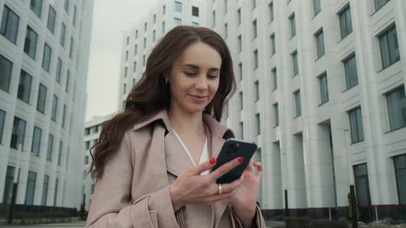 Cheerful young Caucasian woman standing in city street and texting on mobile phone with smile. alt