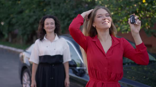 Portrait of Beautiful Young Woman in Red Dress Bragging Car Key in Slow Motion with Blurred Mature alt