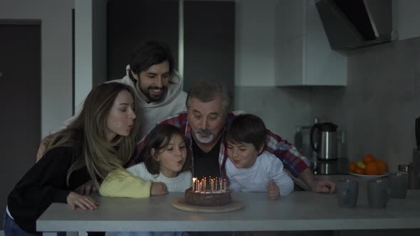 Happy Grandfather Blowing Candles on Birthday Cake Surrounded By His Family alt