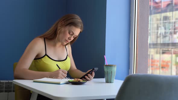 A Young Caucasian Woman Sits in a Yellow Tshirt in a Cafe with Blue Walls and Writes with a Pen in a alt