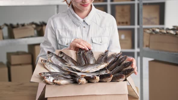 Small Business Charming Female Saleswoman Packing Dried Fish Into Boxes While Sitting on Background alt
