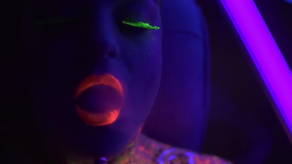 Portrait of a Young Beautiful Woman with Fluorescent Makeup Inhaling Hookah Under UV Black Light alt
