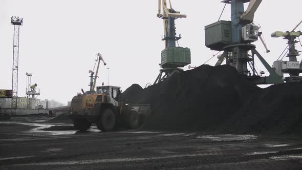 Loader Picks Up Bucket of Coal Against the Backdrop of Seaport with Huge Cranes