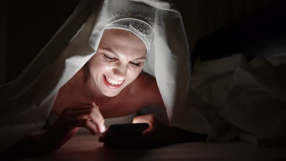 Beautiful Young Woman Using Smart Phone Under Blanket in Bedroom on Bedtime alt