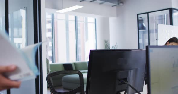 Diverse male and female colleague wearing face masks elbow bumping at desk in busy office alt