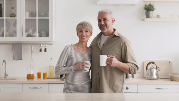 Happy MiddleAged Spouses Holding Coffee Cups Posing Standing In Kitchen alt