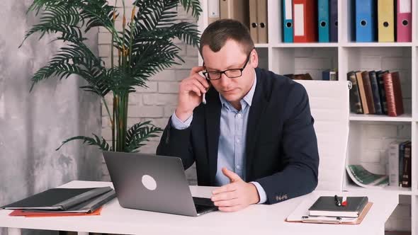Disgruntled Frustrated Businessman in Glasses and Jacket Talking on Mobile Phone Sitting in Office alt