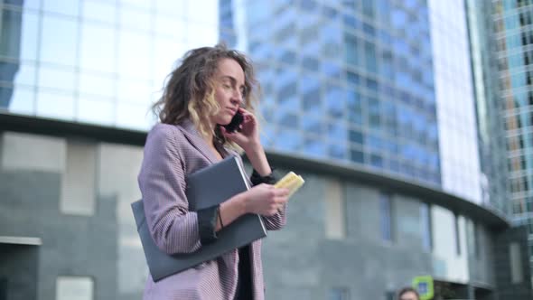 Side view of young business woman eating on the go and talking on mobile phone alt