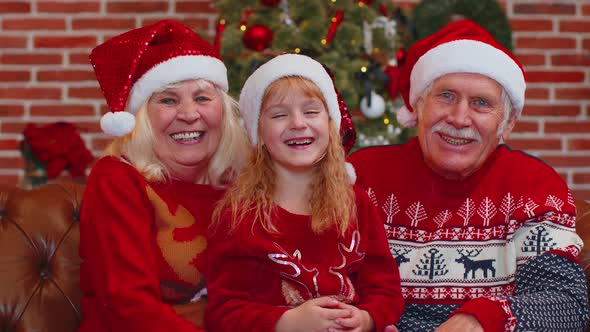 Senior Grandparents with Granddaughter in Santa Hats Laughing Out Loud at Home Near Christmas Tree alt