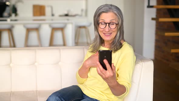Positive Senior Mature Lady in a Yellow Jumper Talking Online on the Phone alt