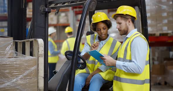 African Female Fork Lift Truck Driver Discussing Checklist with Foreman in Warehouse alt