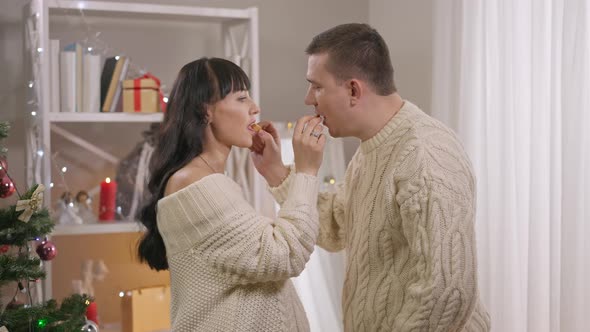 Side View of Romantic Couple Feeding Each Other with Sweet Delicious Candies on Christmas at Home alt
