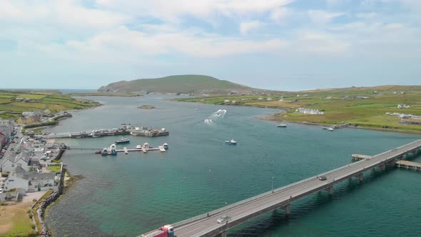 Trucks and cars travel over long bridge onto an island while a selection of boats leave harbor over alt
