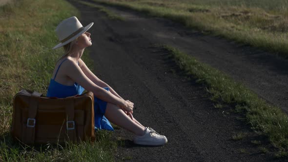 Girl in blue dress with suitcase sit on country road in summer.