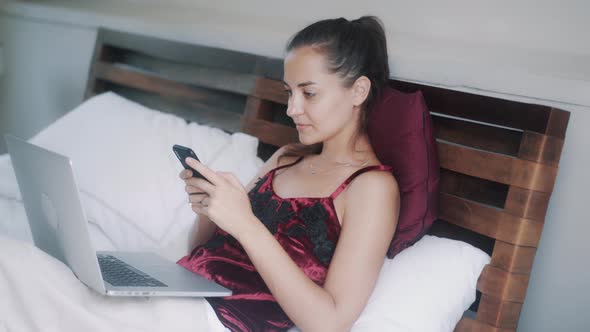 Woman Lies in Bed and Uses Telephone and Laptop for Work at Home During Pandemic alt