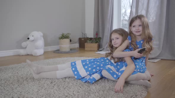 Two Cute Caucasian Girls Sitting on Soft Carpet Indoors with Remote Control, Twin Sisters in Blue alt