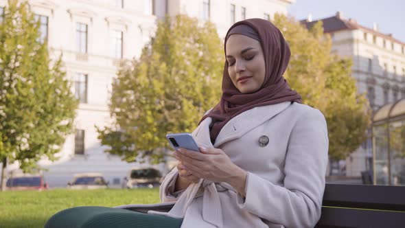A Young Beautiful Muslim Woman Works on a Smartphone with a Smile As She Sits in a Park alt