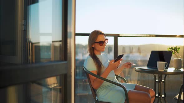 Woman Sitting on the Balcony Against the Backdrop of the Setting Sun and Makes an Online Purchase alt
