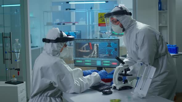Chemist in Ppe Suit Working at Pc While Lab Technician Bringing Her Blood Samples alt