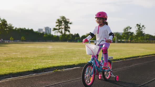 Cute Light Hair Little Girl in Pink Helmet in Elbow and Knee Pads Learns How to Ride a Bicycle alt