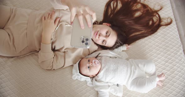 Young Mother Lies On The Bed With The Baby And Takes A Selfie On The Phone