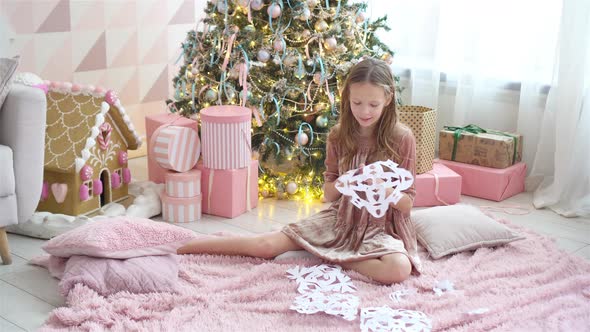 Adorable Little Girl Sitting Near the Tree and Making Paper Snowflakes