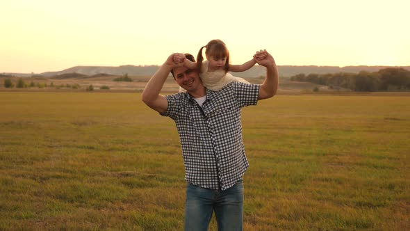 Happy Family Resting in the Park. Father Walks with His Daughter on His Shoulders in Rays of Sunset alt