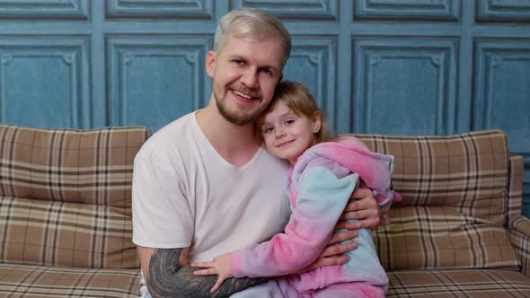 Father and Little Child Daughter Kid in Pajamas Sit on Couch in Room Smiling Looking at Camera alt
