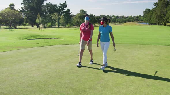 Two caucasian women playing golf wearing face masks walking together alt