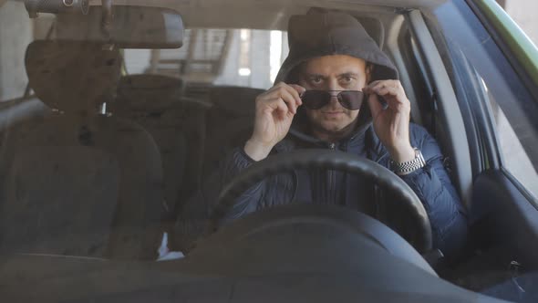 A Man in Black Glasses and a Hoodie at the Wheel of a Car is Secretly Shooting on a Longfocus Lens