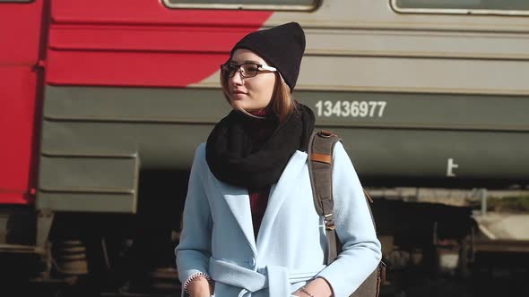 Portrait girl standing on railway platform with backpack  background of train. alt