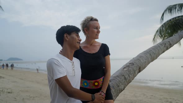 Lovely Cheerful Interracial Couple Chatting While Dating on a Beach Sitting on a Bent Palm and alt