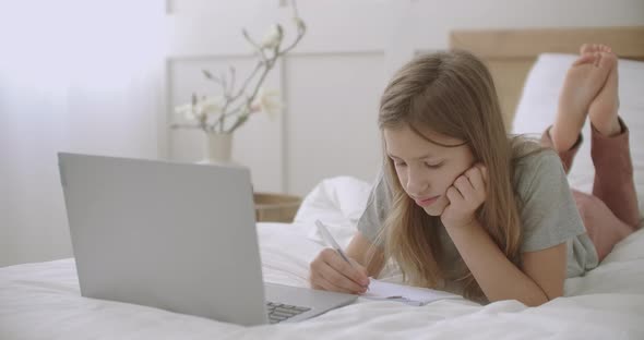 Little Girl Is Drawing and Writing in Copybook, Lying on Bed in Front of Laptop in Her Room, Waiting alt