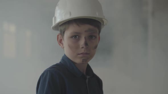 Cute Boy in a Protective Helmet and with Dirty Face Looking at the Camera and Walking in the Smoke alt