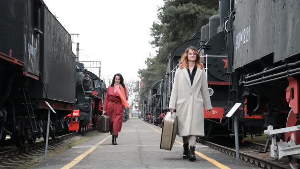 A Young Girl and a Woman Walk on a Railroad Platform with Suitcases alt