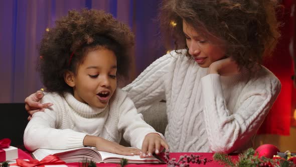 African American Mom and Little Daughter in White Sweaters Read a Book and Cuddle Happily alt