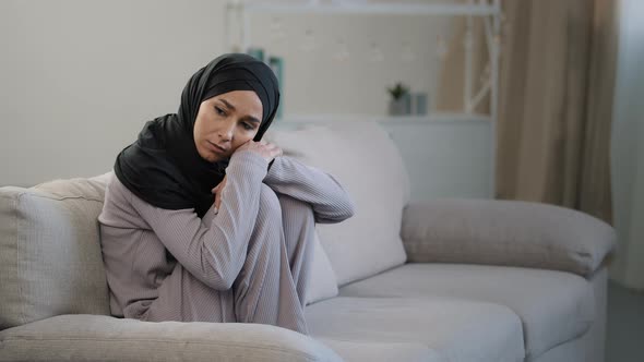 Anxious Sad Young Woman in Black Hijab Sitting on Couch in Living Room Suffering From Psychological alt
