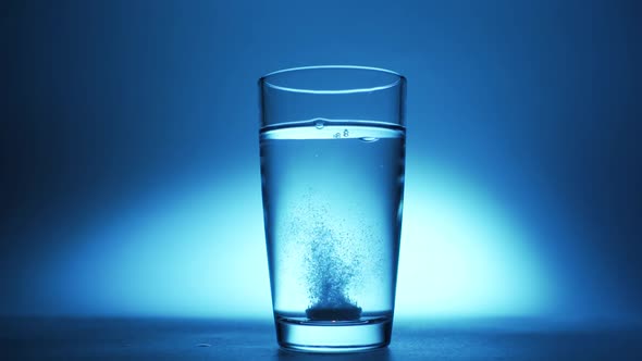 An Effervescent Soluble Tablet Falls Into a Glass of Water. On a Blue Background alt