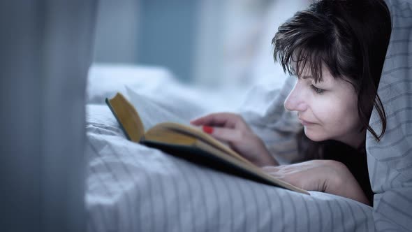 Brunette Happy Woman Covered with a Blanket Lies on the Bed and Reads a Book Wonderful Story About