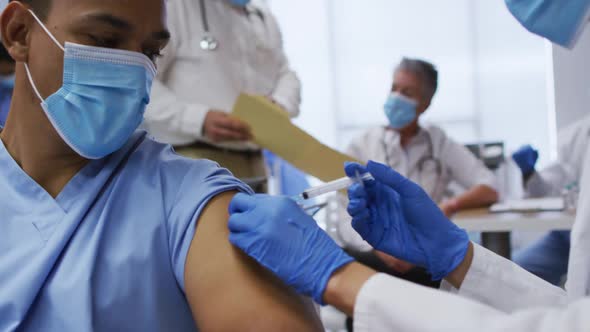 Mixed race female doctor giving covid vaccination to male colleague in hospital, both in face masks alt