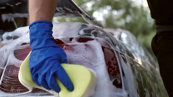 Close Up of a Male Hand in a Blue Glove with a Foam Rubber Sponge for Washing the Headlights alt