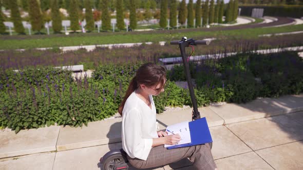 Woman Worker Develops Technology in a Notebook, Sitting on an Electric Scooter in a Beautiful Park