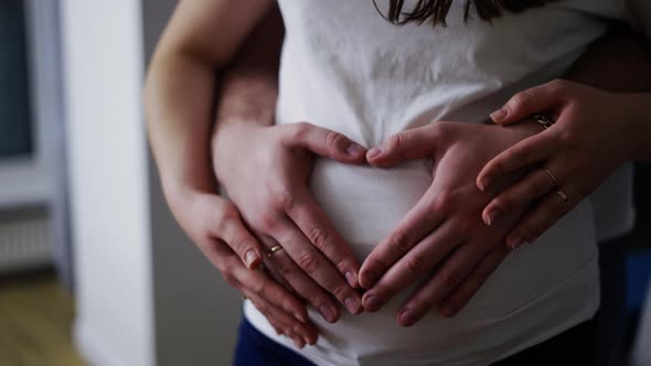 Loving Young Husband and Wife Make Heart Sign Hugging Pregnant Belly at Home alt