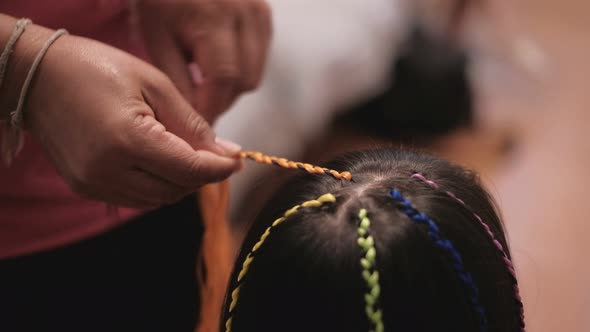 Asian girl is being braided in multi-colored threads by woman's hand. alt