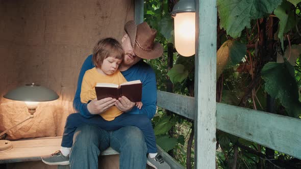 Man in Cowboy Hat Reads Paper Book to Child Sitting on Porch of Village House