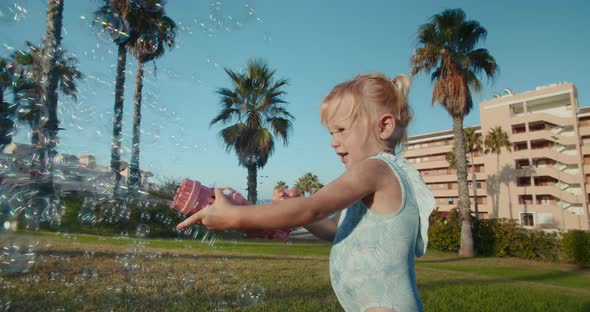 Cute Baby Girl Playing with Bubbles Gun in the Park Standing on Green Grass alt