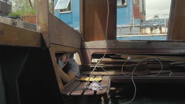 Young man wearing mask painting underside of deck interior wooden boat cabin. alt