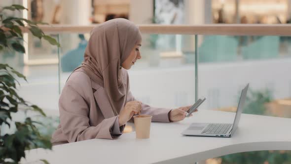 Young Muslim Businesswoman in Hijab Sitting in Office Uses Phone Stirring Coffee in Paper Cup alt