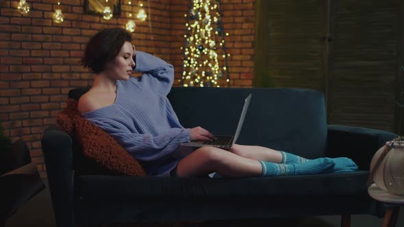 Pretty Girl Relaxing on Sofa in Cozy Room and Uses Laptop alt