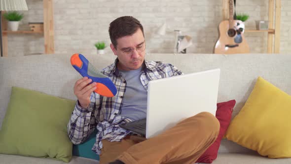 Man Sitting at a Table Uses a Laptop and Holds Orthopedic Insoles in Hand alt